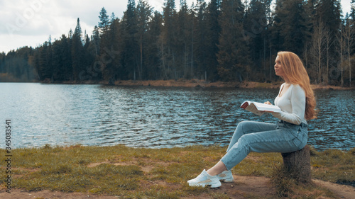 Beautiful young blonde woman sitting and reading book in a park near the lake. Side view