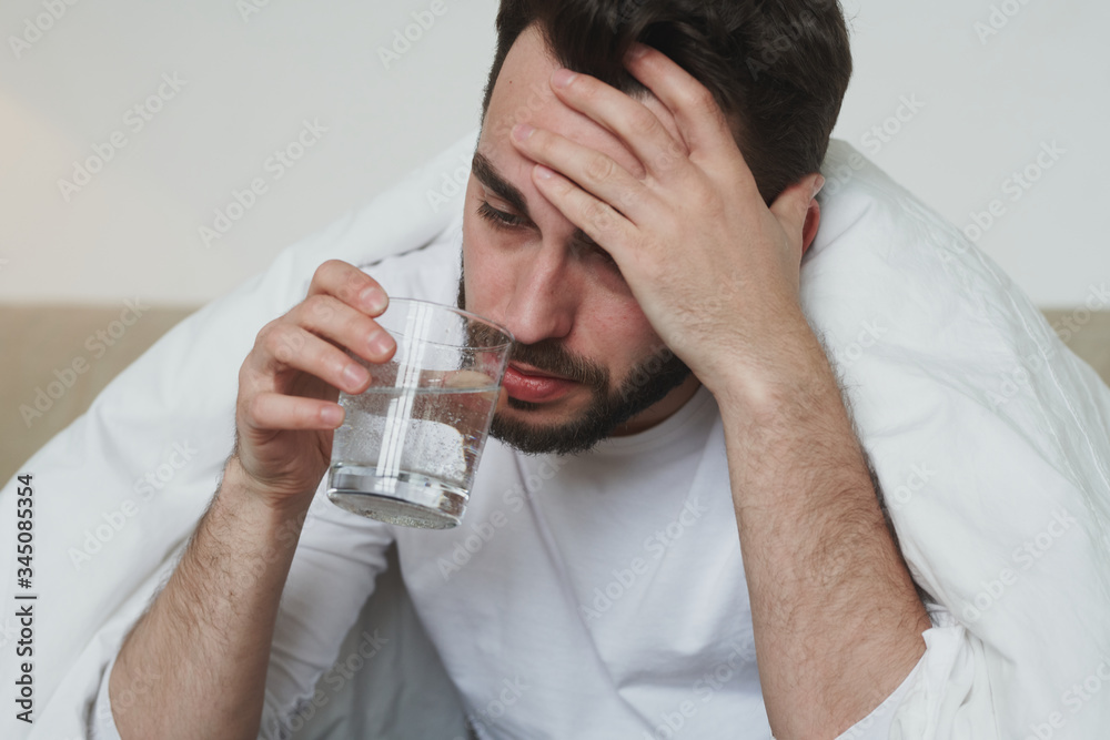Unhappy young man with covid19 or flu touching his forehead and drinking water Unhappy young man with covid19 or flu touching his forehead and drinking water