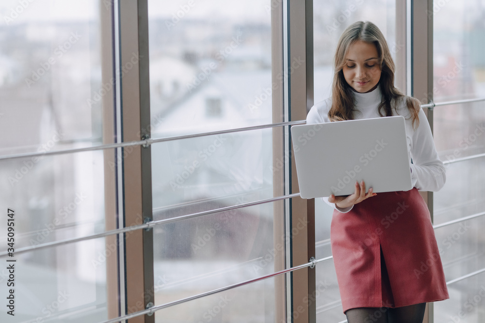 pretty young girl with laptop by the window in office with big windows. remote work concept. freelancer working alone. pretty young girl with laptop by the window in office with big windows. remote work concept. freelancer working alone.