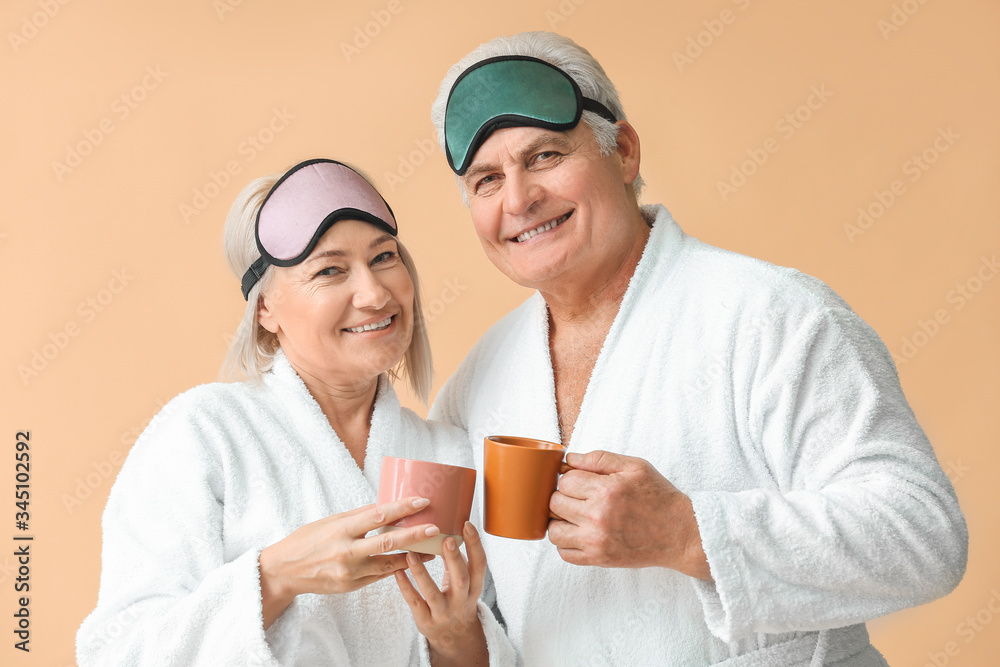Mature couple in bathrobes and with sleep masks drinking tea on color background Mature couple in bathrobes and with sleep masks drinking tea on color background