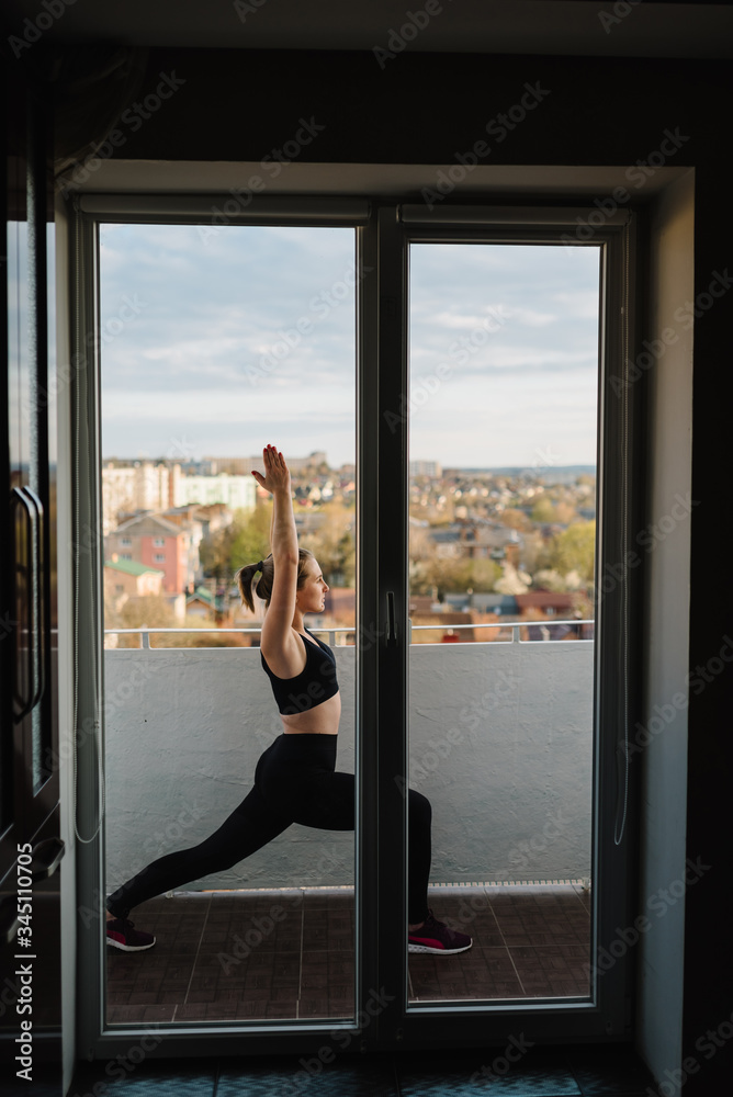 A woman practices yoga on the balcony of her apartment during the Covid-19 lock down. Sport at home. A sports woman is focused on the practice of yoga asanas, an open bright terrace with city views.