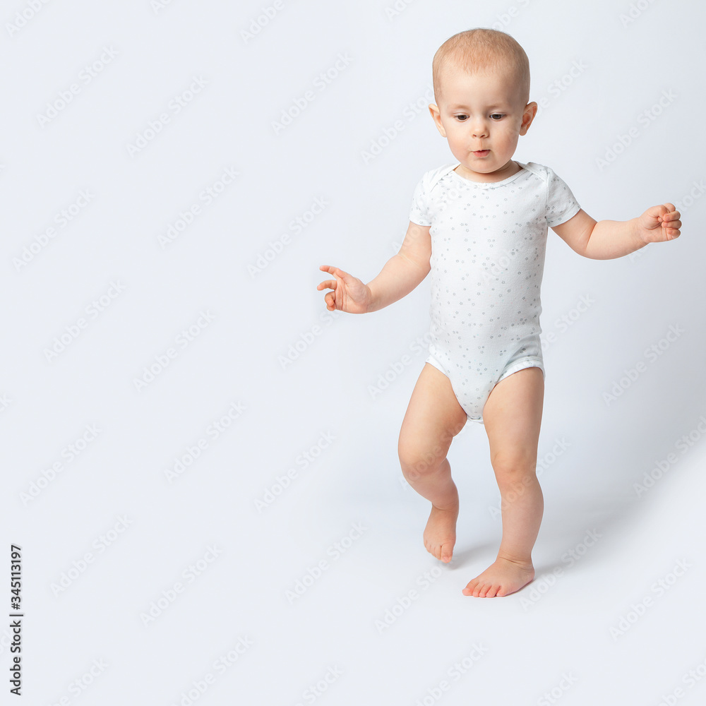 The first steps of the baby. The little girl walks unsteadily, splay-footed, spread her hands to the sides for balance. A child on a white background in a white bodysuit. The first steps of the baby. The little girl walks unsteadily, splay-footed, spread her hands to the sides for balance. A child on a white background in a white bodysuit.