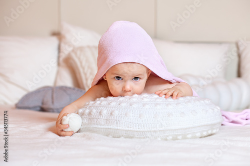 Funny baby after bathing in a bright room on a white bed looks out from behind the pillow. Pink baby towel. Textiles and bedding for children. Family morning at home. The mother and baby.