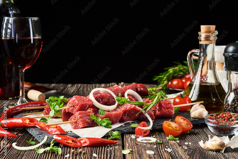Five bamboo skewers with fresh beef meat lie on a stone board on an old wooden table. In the background, various condiments, vegetables, spices and herbs. Free space for text.