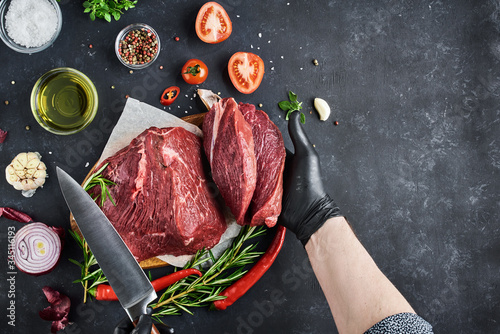 The butcher shows a section of fresh meat tenderloin, which lies on parchment on a dark background. On the table-seasonings, spices, vegetables and herbs. Top view, free space for text. photo