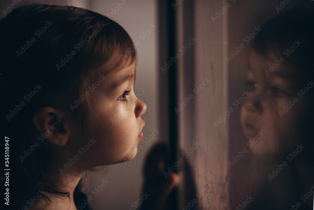 Toddler Caucasian cue girl in looking in the window in self-isolation during covid virus feeling bored