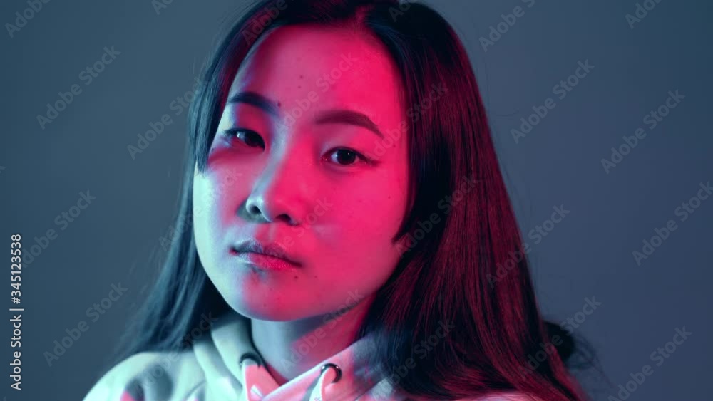 Young pretty girl with asian appearance in neon pink and blue lights at night. Serious trendy hipster teenager looking to camera Young pretty girl with asian appearance in neon pink and blue lights at night. Serious trendy hipster teenager looking to camera