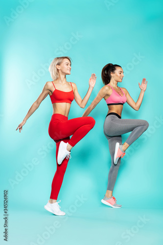 Sporty girls. Side view of two young beautiful women in sportswear jumping in studio against blue background. Sport and fitness