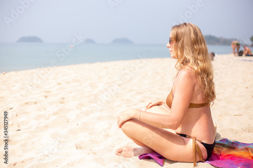 European woman in a swimsuit, with long blonde hair, sitting in a lotus position on the sand by the sea, side view