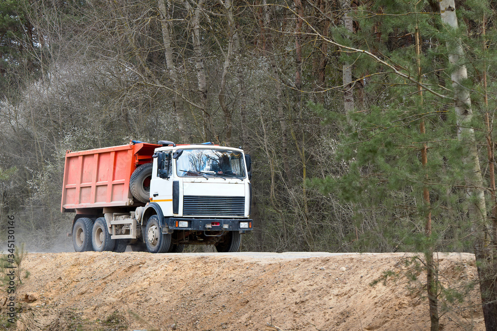 Heavy laden multi-ton truck with white cab and orange body rides along dusty country road in with spring forest background leaving behind clouds of dust.