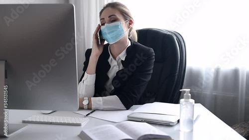 Work during self-isolation and quarantine. Young woman employee in a medical facial mask works in the office at the computer and talking mobile phone. Coronavirus Pandemic Covid 19.