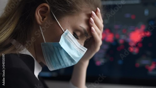 Side view close-up portrait of a young woman in disposable medical facial mask holding her head and looking at the epidemic map of the coronavirus Covid 19 pandemic on a computer