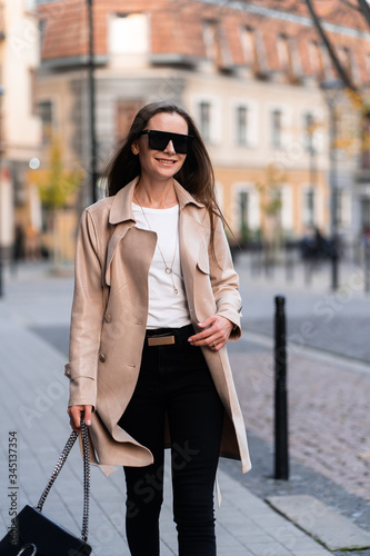 Obraz na płótnie Outdoor photo of brunette lady walking on street background in autumn day.Fashion street style portrait. wearing dark casual trousers, white sweater,creamy coat and sunglasses.Fashion concept. w salonie