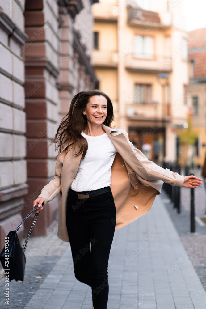 Outdoor photo of brunette lady walking on street background in autumn day.Fashion street style portrait. wearing dark casual trousers, white sweater and creamy coat.Fashion concept.