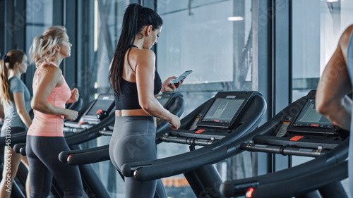 Athletic Sports Woman in Gym Puts on Wireless Headphones, Turns on Sport Music Playlist with Smartphone and Starts Running on Treadmill. In Background Fit Athletes Training.