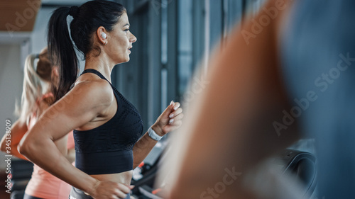 Fototapeta Naklejka Na Ścianę i Meble -  Beautiful Athletic Sports Woman Wearing Wireless Headphones, Listens to a Podcast or Sport Music Playlist while Running on a Treadmill. In Background Fit Athletes Training in the Gym