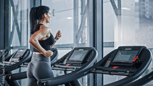Beautiful Athletic Sports Woman Wearing Wireless Headphones, Listens to Podcast or Sport Music Playlist while Running on a Treadmill. Energetic Female Athlete Training in the Gym Alone. Side Back View