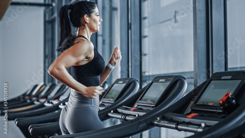 Beautiful Athletic Sports Woman Wearing Wireless Headphones Listens to Podcast or Sport Music Playlist while Running on Treadmill. Energetic Female Athlete Training in Gym Alone.