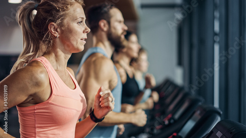 Fit Athletic Woman Running on the Treadmill, Doing Her Fitness Exercise. Muscular Women and Men Actively Training in the Modern Gym. Sports People Workout in Fitness Club. Side View