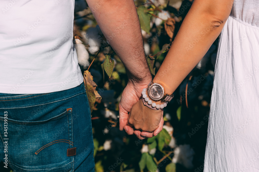 A couple holding hands in cotton field. Woman in white dress and man in blue jeans and white T-shirt. Girl and boy holding hands. Couple on valentines day. Clock and decoration on woman's hand
