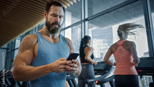 Muscular Heavyweight Champion Walks Through Gym, Uses Smartphone for Social Media and Conducting Business Affairs. In the Background Sports People Running on Treadmills