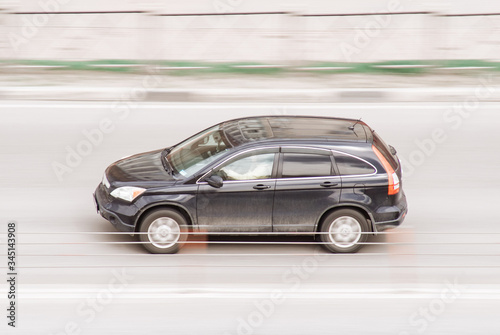 Fast moving black car on the city road. SUV in motion side view. Vehicle driving along the street in city with blurred background. Compliance with speed limits on road