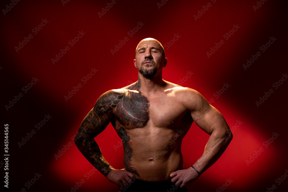 Muscular man with tattoo and closed eyes posing on red background with naked torso, hands on the sides. Muscular body, studio shot
