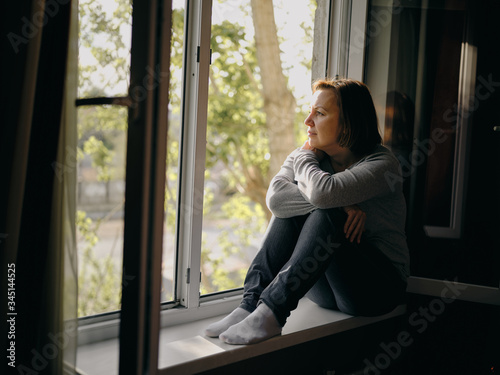 A beautiful young woman is sad sitting on a window in her house. Quarantine and self-isolation due to the coronavirus pandemic.