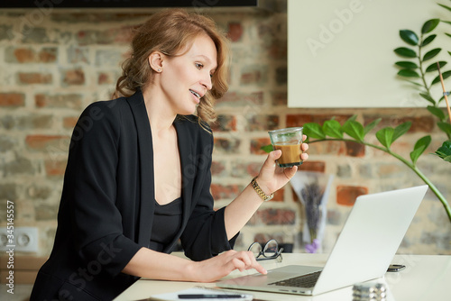 A happy woman with braces working remotely on a laptop in a kitchen. A lady with a coffee discussing a project with colleagues at a video conference at home. A teacher preparing for an online lecture