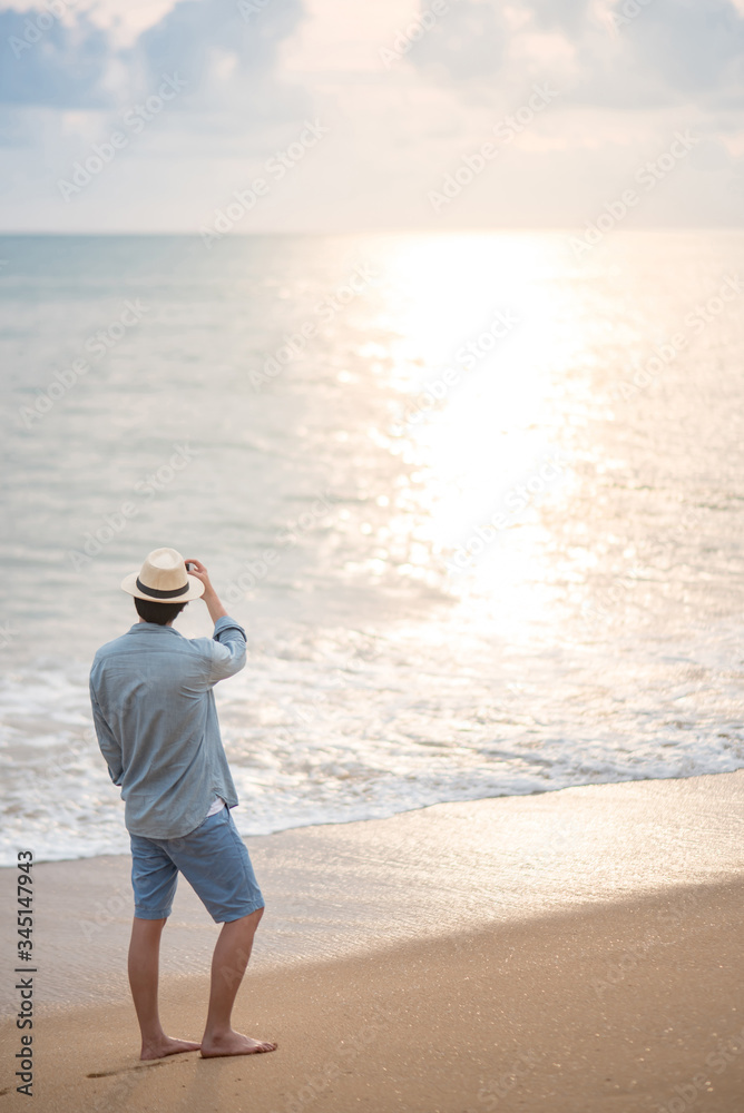 Asian man tourist wearing hat standing on tropical island beach enjoy looking at sunset. Relaxing holiday or vacation travel in summer season. Summertime trip concept