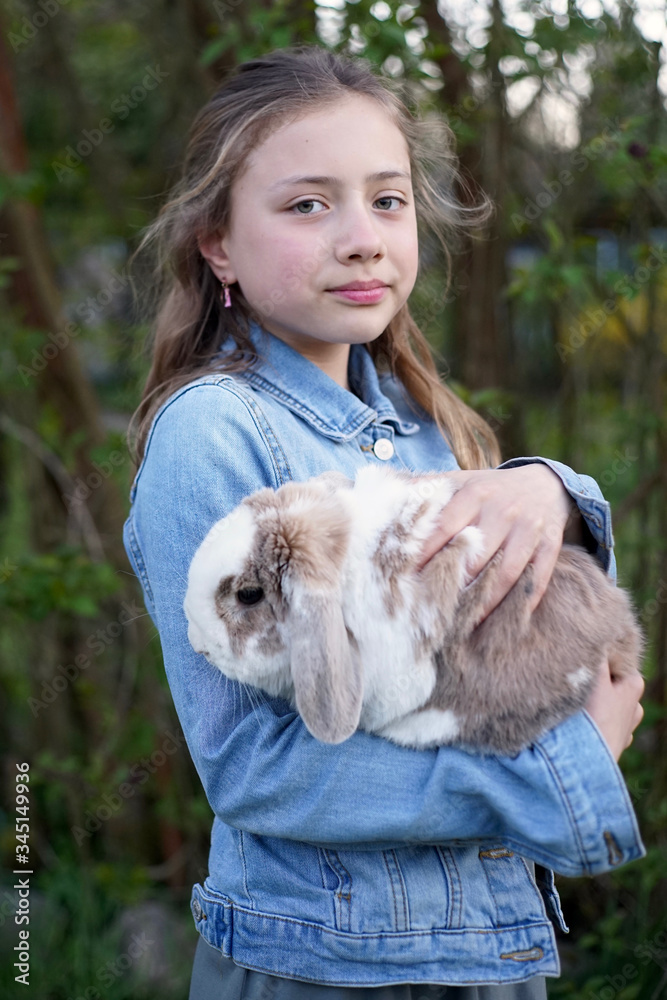 Portrait of a young blonde girl holding gently a pet bunny, rabbit in her arms outside in the garden.    