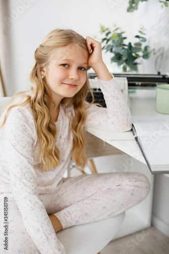 Home schooling girl weared in sleepwear. Child girl in pajamas sitting at the desk. Blonde girl doing homework While Schools Are Closed. Home time. Learning Remotely. Girl Does Schoolwork At Home