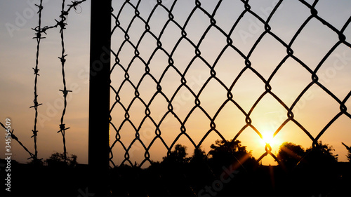 Selective focus on silhouette blurred on mesh fence and barbed wire at dark sunset.