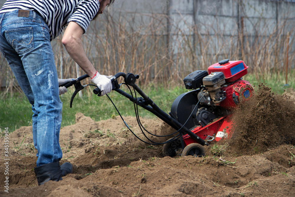 Obraz premium Man working in the garden with garden tiller. tractor cultivating and loosens soil field