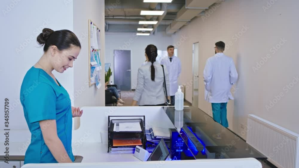 In the modern office at the reception beautiful young nurse meeting patient at the enter of the hospital at says where she have to go other doctors and stuff walking around the reception