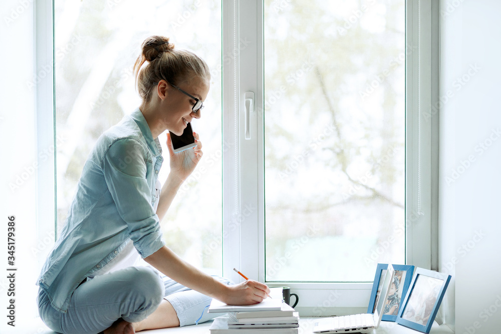 Young woman working from home office. A freelancer uses a laptop, phone, internet and notepad. Workplace on the windowsill. The concept of female business, career, communication, online learning Young woman working from home office. A freelancer uses a laptop, phone, internet and notepad. Workplace on the windowsill. The concept of female business, career, communication, online learning