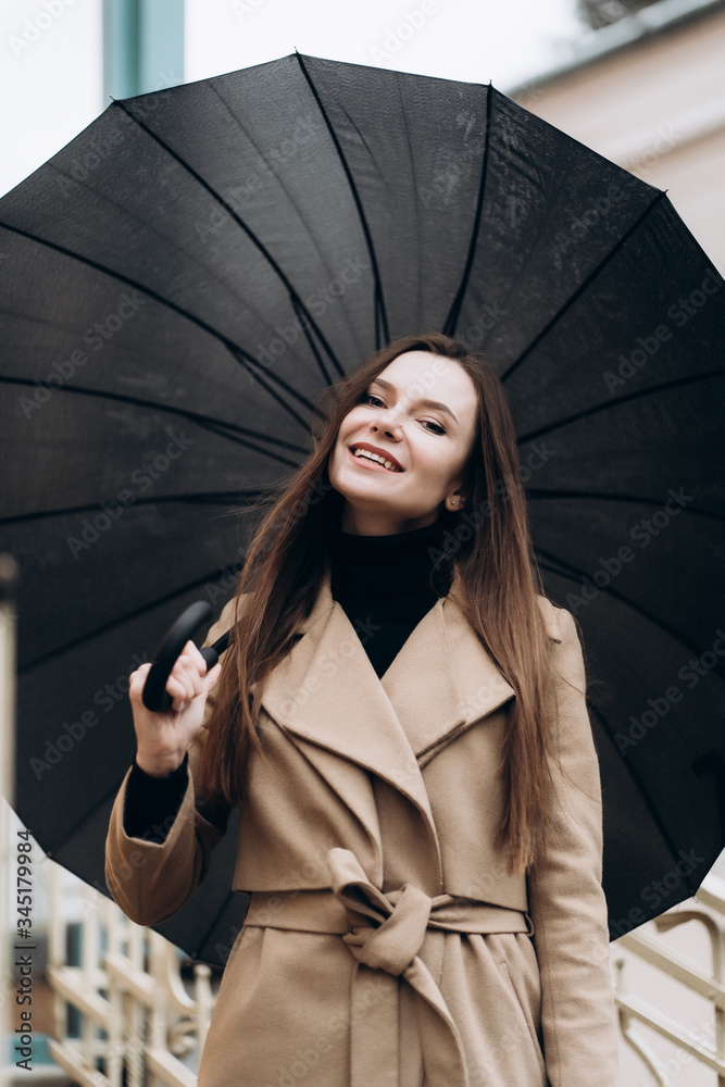 Outdoor photo of brunette lady posing with black umbrella in rainy autumn day close up.Fashion street style portrait. wearing dark casual trousers, white sweater and creamy coat.Fashion concept.