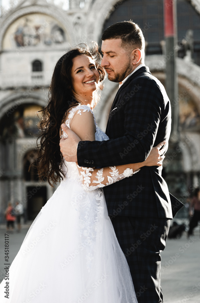 Naklejka premium happy bride and groom cuddling while walking around the old city of venice,groom of Georgian appearance hugs a bride of European appearance,european wedding,lovely wedding couple look at each other
