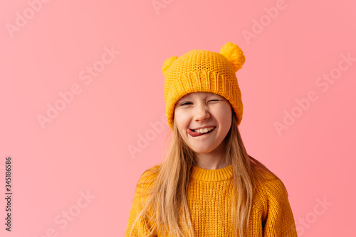 Happy little girl dressed for spring wearing yellow hat on pink background sticking out her tongue