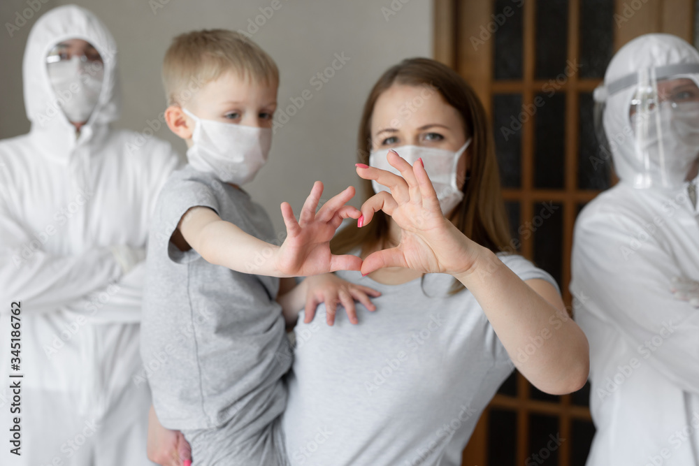 Mother and son show a heart sign with their hands against the background of infectious disease workers in white protective suits during the coronavirus epidemic. Covid-19