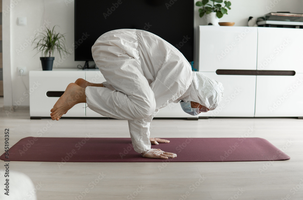 Young man in medical mask and in protective suit is practicing yoga on a mat at home. Stay home during COVID-19 quarantine concept. Yoga at home online. Coronavirus pandemic. Healthy lifestyle.