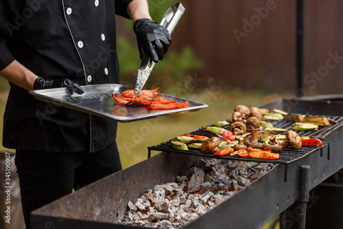 Cook in black suit remove hot pepper from grates. A lot of vegetables lie on the grill. Catering food preparation for outdoor party. Barbeque for big company