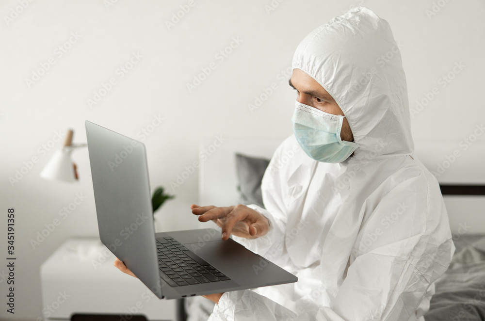Man in protective white suit and medical mask is working from home on a bed with laptop because of coronavirus epidemic. Remote work during pandemic. Stay home during COVID-19 quarantine concept. Man in protective white suit and medical mask is working from home on a bed with laptop because of coronavirus epidemic. Remote work during pandemic. Stay home during COVID-19 quarantine concept.
