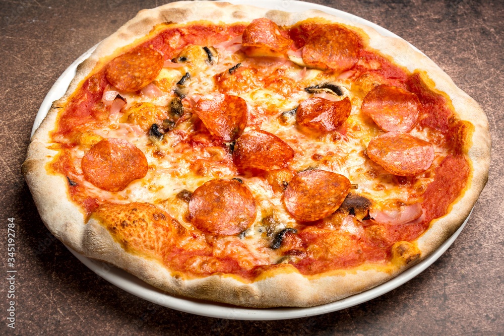 Pizza with salami, ham, mushrooms, tomato sauce, thin dough, on plate, brown background.