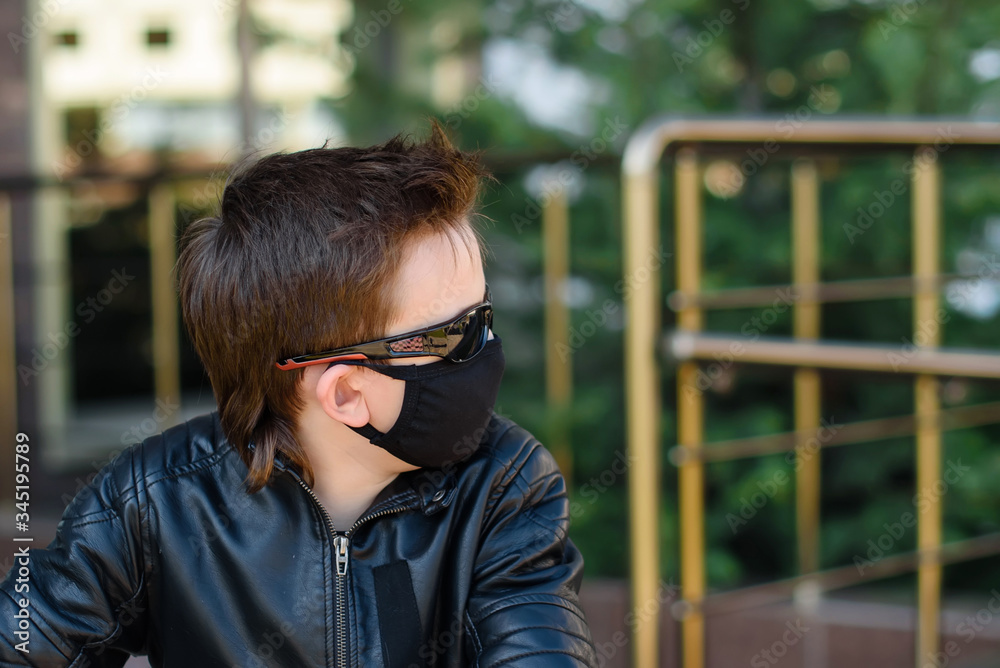 Boy in a leather jacket wearing a black hygyenic medical mask outdoors in the yard in black sunglasses, looking forward. View from the side. Cancelled vacation, bad mood due to coronavirus covid-19 Boy in a leather jacket wearing a black hygyenic medical mask outdoors in the yard in black sunglasses, looking forward. View from the side. Cancelled vacation, bad mood due to coronavirus covid-19