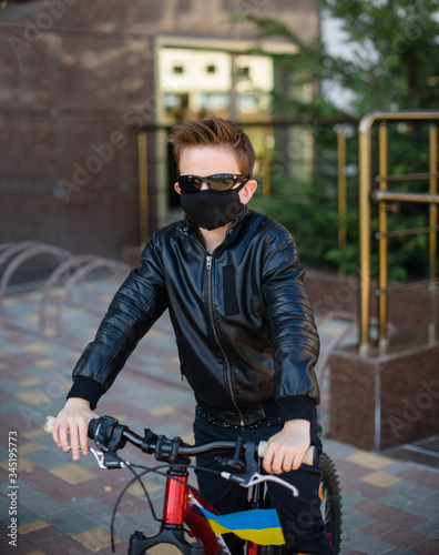 Boy in a leather jacket wearing a black hygyenic medical mask outdoors in the yard in black sunglasses, sitting on a bike with ukranian flag.