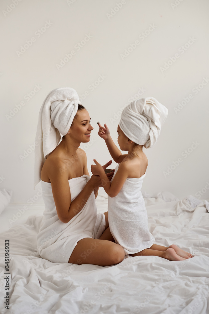 A young caucasian mother and little daughter with wrapped hair in white bath towels apply a clay mask on the mother faces
