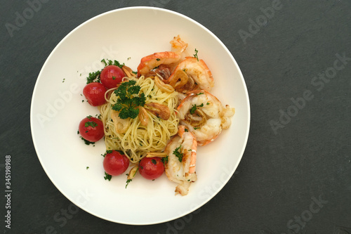 High angle shot of spaghetti with fried scampi, garlic slices, cherry tomatoes decorated with chops parsley on white round plate and dark grey black slate slab board background.