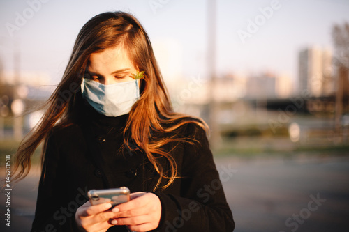 Portrait of sad teenage girl using smart phone wearing protective face medical mask standing outdoors on city street
