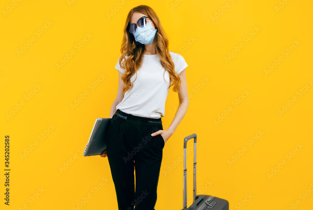 business woman with a protective medical mask on her face, with a laptop and a suitcase on a yellow background. The concept of travel, quarantine, and coronavirus business woman with a protective medical mask on her face, with a laptop and a suitcase on a yellow background. The concept of travel, quarantine, and coronavirus